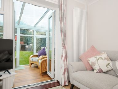 A living room with a gray sofa and floral cushions next to glass doors leading to a conservatory with wicker chairs at St Albans in Boscastle