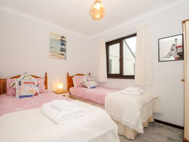 A bedroom with two single beds with pink bedding two folded towels on each bed a wooden nightstand with a lamp a window with white curtains and wall art at St Albans in Boscastle