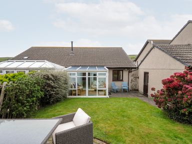 A backyard with grass and garden furniture in front of a house with a glass conservatory at St Albans in Boscastle