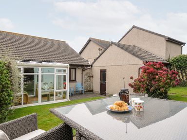 A garden with a glass table set with croissants and mugs next to a conservatory and two houses at St Albans in Boscastle