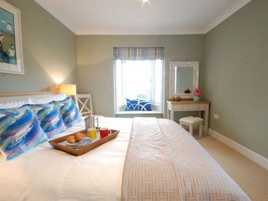 A bedroom with a bed pillows a tray with mugs and bagels a window with seating and cushions and a dressing table with mirror at Crabbe Corner in Aldeburgh