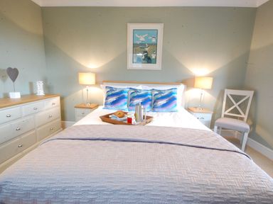 A bedroom with a bed with colorful pillows and breakfast tray two bedside tables with lamps a white chair and a dresser at Crabbe Corner in Aldeburgh