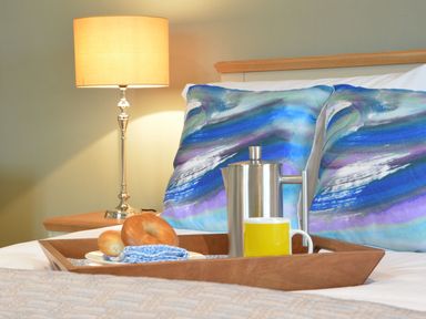 A bed with a wooden tray holding a french press, a yellow mug, bagels, and a blue napkin beside a bedside lamp at Crabbe Corner in Aldeburgh