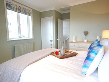 A bedroom with a bed with blue and purple pillows a tray with breakfast on the bed a window with striped blinds and a dresser with decorations at Crabbe Corner in Aldeburgh