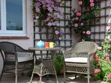 An outdoor seating area with a wicker table and two wicker chairs near a wall with climbing pink and purple flowers at Crabbe Corner in Aldeburgh