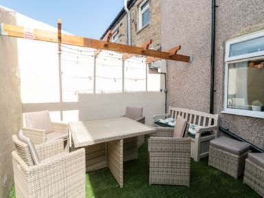 A small outdoor patio with a wicker table surrounded by four wicker chairs and a bench with cushions at Bonnies Retreat in Amble