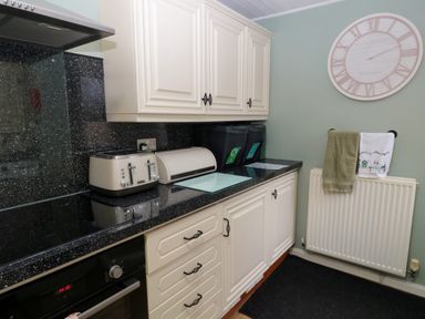 A kitchen with black countertop white cabinets a toaster a bread bin waste bins a wall clock and towels hanging over a radiator at Bonnies Retreat in Amble