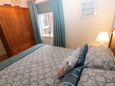 A bedroom with a wooden wardrobe a bed with patterned bedding and pillows a window with blue curtains and a lamp on a bedside table at Bonnies Retreat in Amble