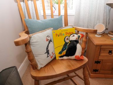 A wooden chair with a puffin cushion a children's book titled Puffin Peter and a puffin plush toy next to a wooden nightstand with a small round mirror at Bonnies Retreat in Amble