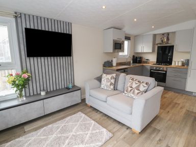 A living room with a grey sofa, a wall-mounted TV, flowers on a cabinet, and a kitchen with appliances at 24 Sherwood in Carnforth