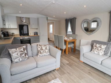A living room with grey sofas and patterned cushions next to a dining table with grey chairs and a kitchen area at 24 Sherwood in Carnforth