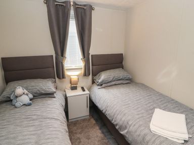 A bedroom with two single beds separated by a nightstand with a lamp and a window with curtains at 24 Sherwood in Carnforth