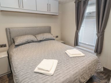 A bedroom with a double bed with striped bedding two folded towels on the bed bedside tables overhead cupboards and a window with curtains at 24 Sherwood in Carnforth