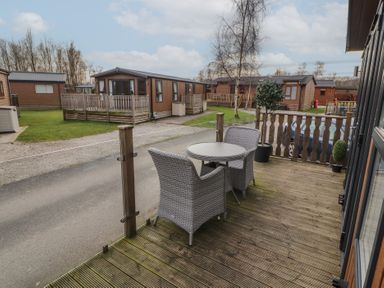 A wooden deck with two wicker chairs and a round table overlooking a road and several brown wooden cabins at 24 Sherwood in Carnforth