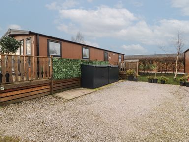 A gravel driveway with a wooden decked area and a brown mobile home with surrounding plants at 24 Sherwood in Carnforth