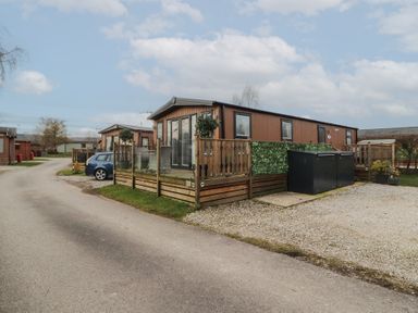 A wooden mobile home with a fenced deck and outdoor seating near a gravel parking area at 24 Sherwood in Carnforth