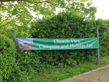 A green banner sign attached to wooden posts with trees and bushes behind it at Church View in Atwick