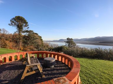 An outdoor seating area with a wooden bench and round table overlooking a river with hills and trees at Bryn Mel Manor Straits View in Menai Bridge