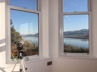 A corner with two windows overlooking a body of water and hills with a telescope on a tripod at Bryn Mel Manor Straits View in Menai Bridge