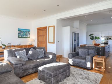 A living room with gray sofas and an ottoman next to a kitchen with white cabinets and a wooden door at Bryn Mel Manor Straits View in Menai Bridge
