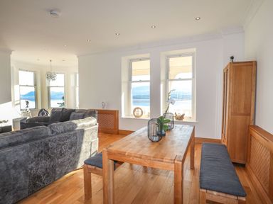 A dining table with bench seating and a living room with gray sofas near windows at Bryn Mel Manor Straits View Menai Bridge