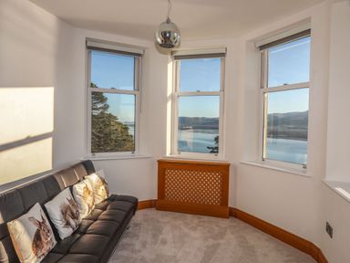 A sitting area with a black sofa with animal-themed cushions and three windows showing a water view at Bryn Mel Manor Straits View Menai Bridge