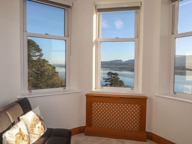 A corner of a room with three windows showing a view of trees and a body of water with hills beyond and a black sofa with animal print cushions Bryn Mel Manor Straits View Menai Bridge