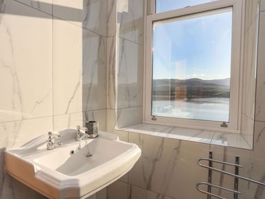 A bathroom sink with taps next to a window overlooking a body of water and hills at Bryn Mel Manor Straits View in Menai Bridge