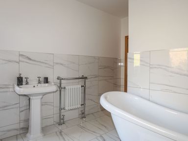 A bathroom with a pedestal sink, a white freestanding bathtub, a towel radiator, and marble wall tiles at Bryn Mel Manor Straits View in Menai Bridge