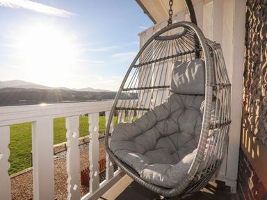 A hanging cushioned chair on a balcony with white railing overlooking green grass and distant hills at Bryn Mel Manor Straits View in Menai Bridge