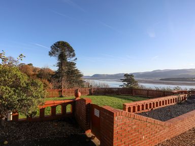 A garden with a brick wall and wooden fence overlooking a body of water and distant hills at Bryn Mel Manor Straits View in Menai Bridge