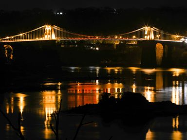 A suspension bridge lit up at night with its lights reflecting on the water at Bryn Mel Manor Straits View in Menai Bridge