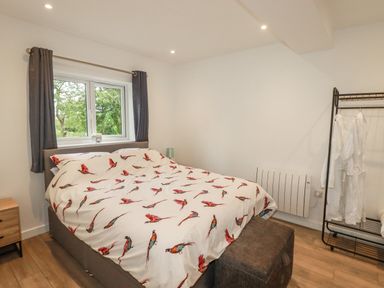 A bedroom with a bed with bird printed bedding a window with curtains a small bedside table a lamp a stool and a garment rack with robes at Pheasant View in Penelewey near Carnon Downs