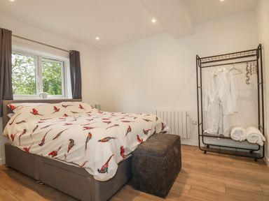 A bedroom with a bed covered in bird patterned bedding a window with curtains a dark ottoman a clothing rack with white robes and towels at Pheasant View in Penelewey near Carnon Downs