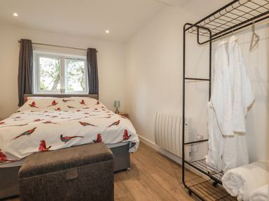 A bedroom with a bed covered in bird patterned bedding a small lamp a window with curtains a standing rack holding white robes and towels at Pheasant View in Penelewey near Carnon Downs