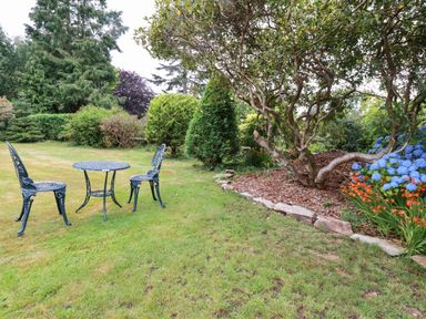 A garden with a metal table and two chairs on grass near trees and flower beds at Pheasant View in Penelewey near Carnon Downs