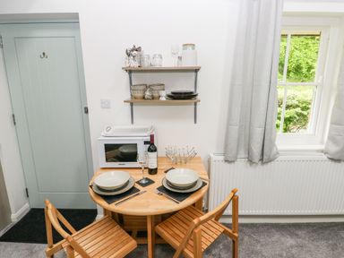 A small dining area with a round wooden table set for two with dishes and wine next to a microwave under shelves with dishes near a window with grey curtains at Ty Rhydd in Newport Pembrokeshire