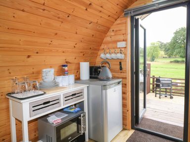 A small kitchen area with a refrigerator microwave kettle plates bowls glasses and mugs with an open door showing a barbecue outside The Pod at Bank House Farm The Blythe near Hixon
