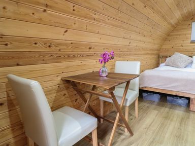 A wooden table with a vase of flowers and two white chairs next to a bed with cushions in a wooden room at The Pod at Bank House Farm The Blythe near Hixon