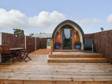 A wooden deck with outdoor chairs and table leading to a small wooden pod with potted plants at The Pod at Bank House Farm in The Blythe near Hixon