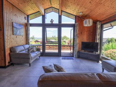 A living room with leather sofas a TV and large windows overlooking a garden at Greengarth in Yanwath near Penrith