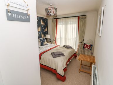 A bedroom with a bed covered in beige and red bedding with folded towels a wooden side table a transparent chair with a cushion and curtains at Yacht Haven View in Neyland