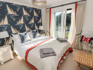 A bedroom with a double bed two pillows patterned with sailboats bedside tables with lamps curtains on sliding glass door and a chair with a cushion at Yacht Haven View in Neyland