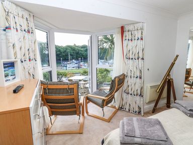 A room with two wooden chairs with striped cushions facing a glass door overlooking a marina with boats and trees with curtains on the windows and a telescope on a tripod in Neyland