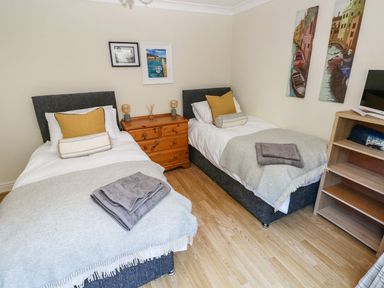 A bedroom with two single beds with gray headboards and blankets a wooden chest of drawers between the beds and a wooden shelving unit with a television on top at Yacht Haven View in Neyland