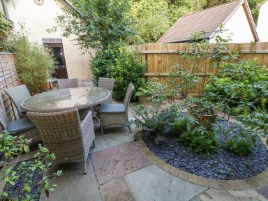 An outdoor patio area with a glass round table and six wicker chairs surrounded by plants and a wooden fence at Yacht Haven View in Neyland