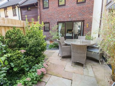 A small backyard patio with a round glass table and six wicker chairs surrounded by plants at Yacht Haven View in Neyland