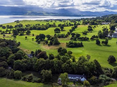 An aerial view of green fields with scattered trees and some houses near a body of water with mountains in the background at Ty Bach - Beaumaris in Beaumaris