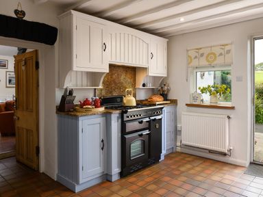 A kitchen with a black stove blue and white cabinets flowers on the windowsill and a wooden door open at Ty Bach - Beaumaris in Beaumaris
