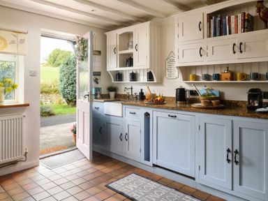 A kitchen with blue lower cabinets white upper cabinets countertop appliances and an open door showing outside at Ty Bach - Beaumaris in Beaumaris